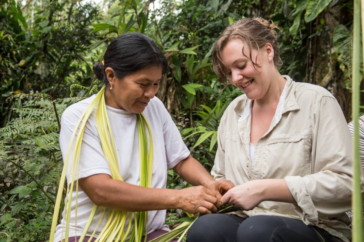 A smiling local woman teaches a visitor how to weave a basket with leaves. 