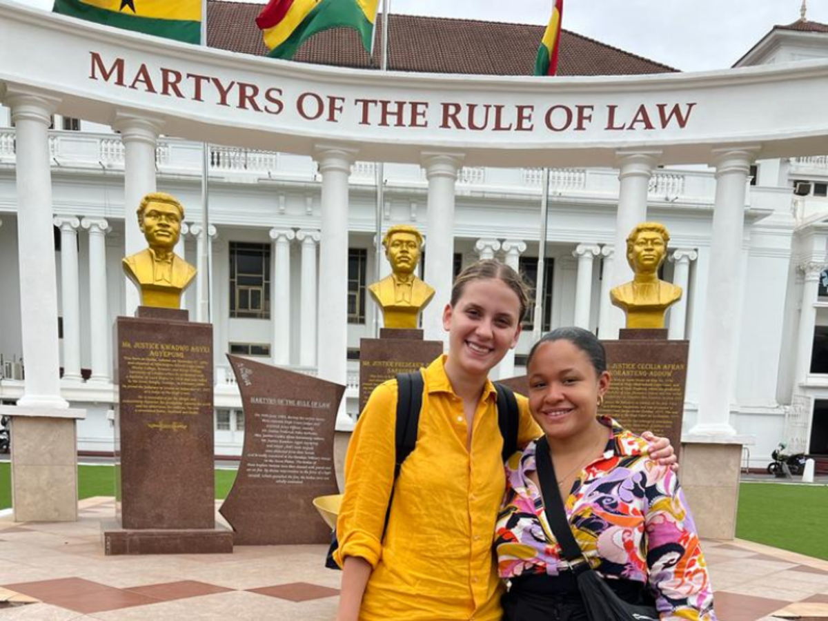 A volunteer and a member of staff stand smiling in front of a monument which reads 'Martyrs of the Rule of Law' - with golden busts, white pillars and Ghanaian flags. 