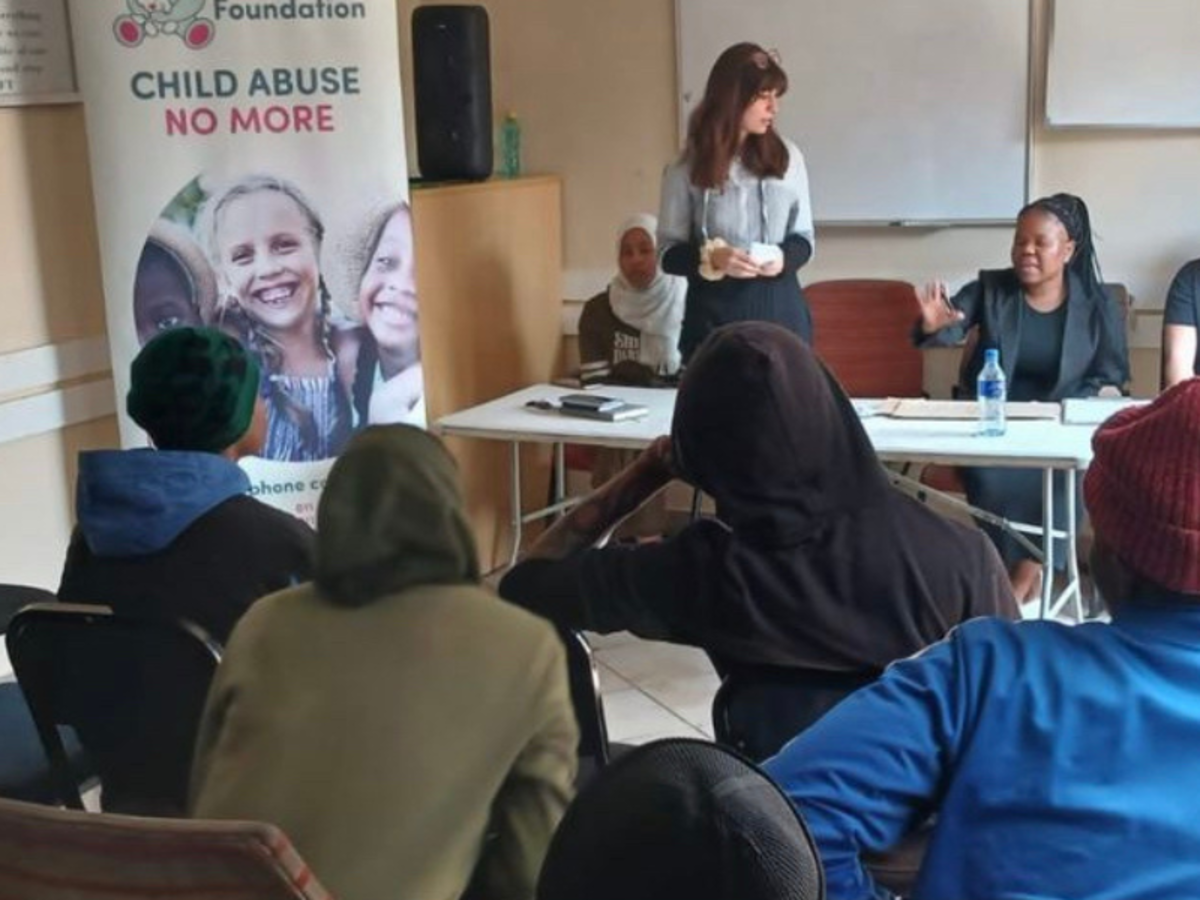 A volunteer stands and addresses a room full of people alongside a member of staff. They sit at a table next to a banner that reads "Child Abuse No More"