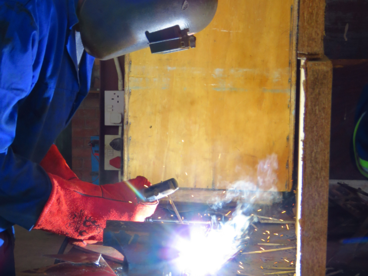 A welder in protective gear, including a helmet and red gloves, works with bright sparks and smoke. The background is a wooden surface, creating a focused, industrious atmosphere.