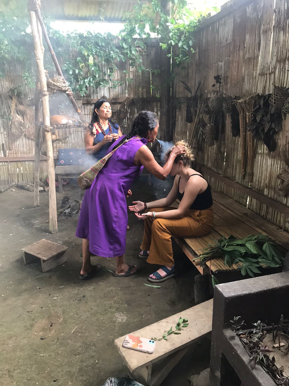 A woman in traditional clothes blows smoke on another's head. 