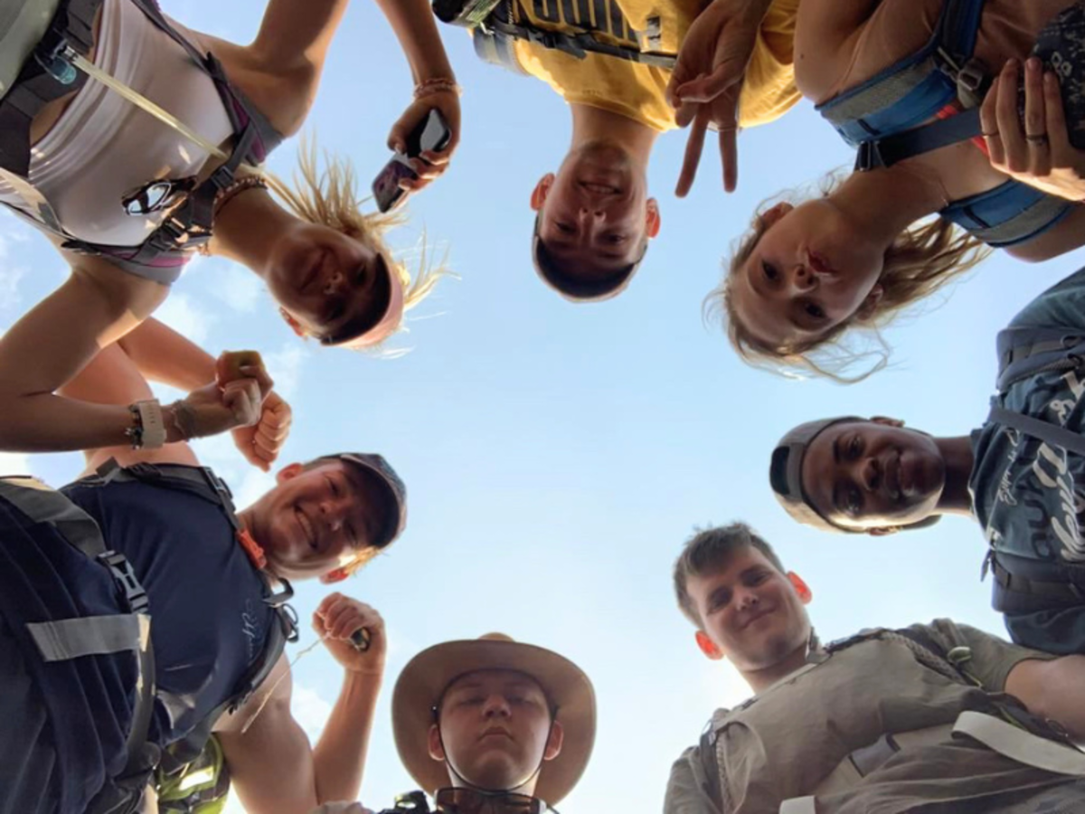 Eight friends form a circle, smiling and showing peace signs, against a clear blue sky. They appear joyful and ready for an adventure.