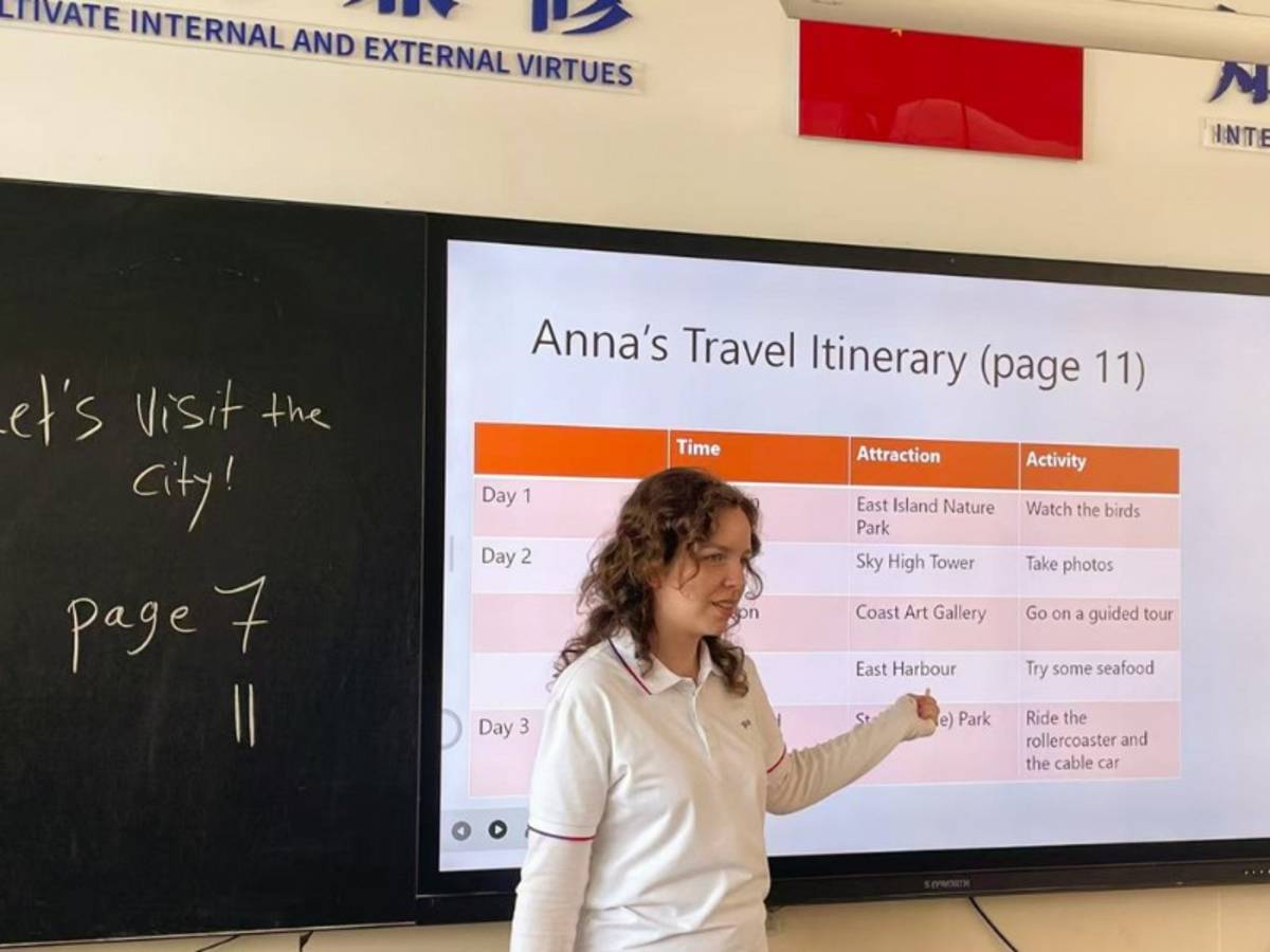 Female teacher points to interactive white board which shows 'Anna's Travel Itinerary'