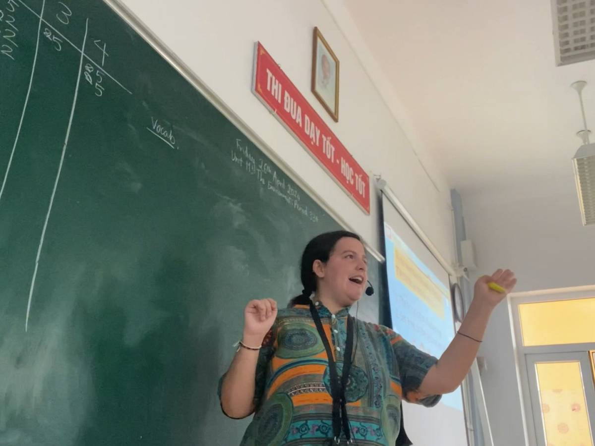 Female TEFL teacher standing in front of chalk board, wearing head microphone and gesturing with arm.