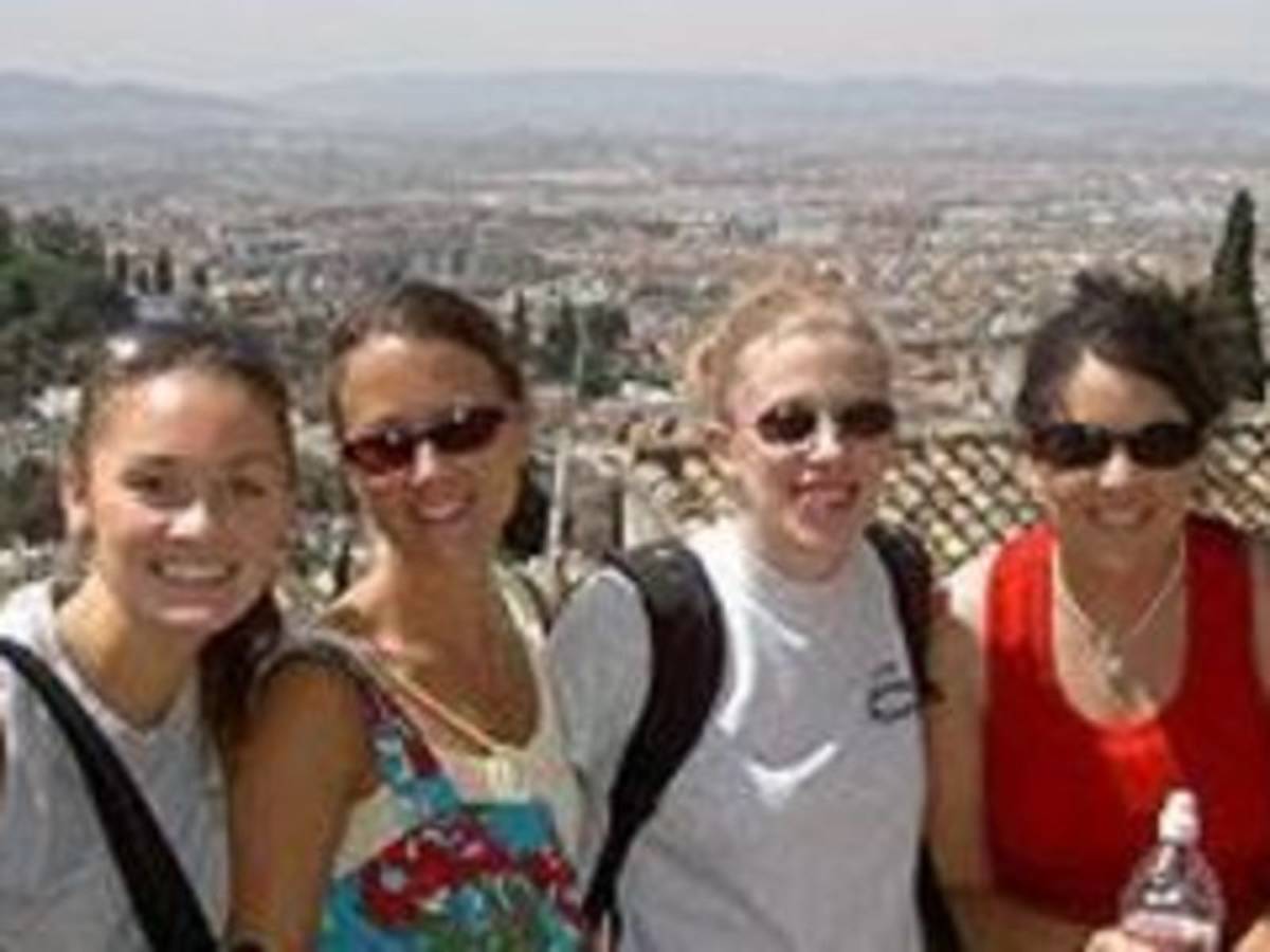 Four women smiling and posing for a picture on a hill, enjoying the beautiful scenery.