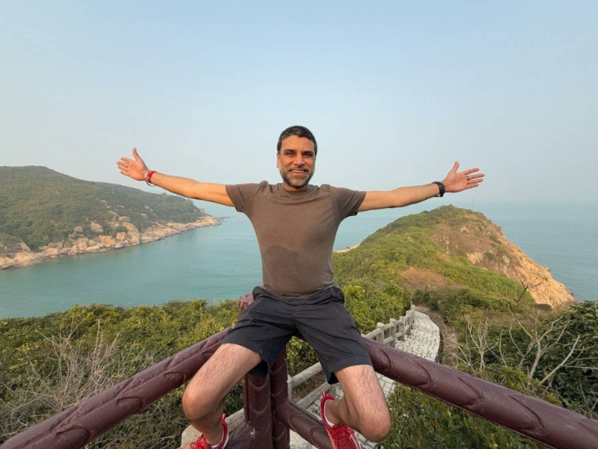 Man sitting on a wooden railing with arms wide open, smiling at a scenic coastal viewpoint with hills and sea in the background.
