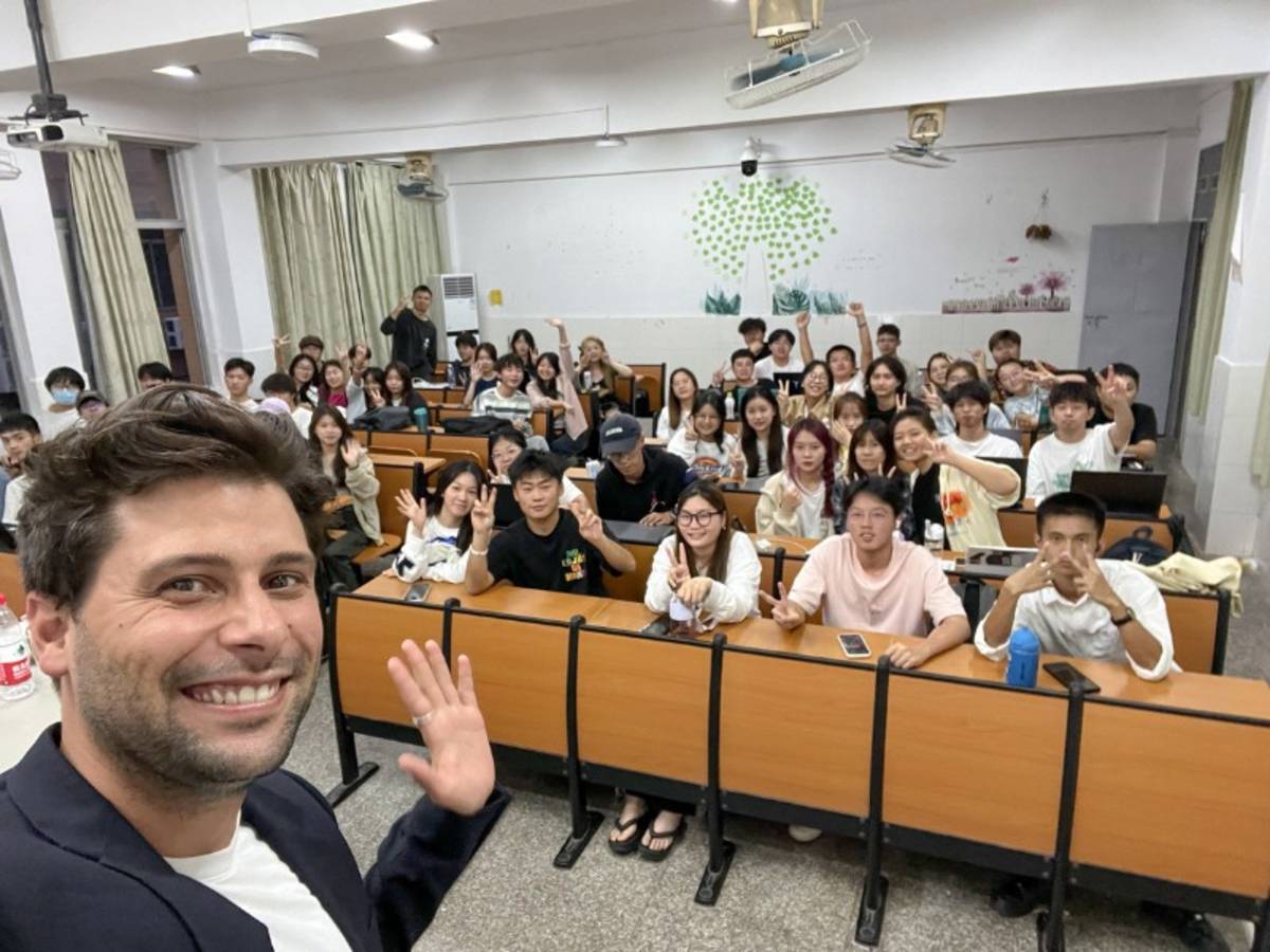 Man smiles and waves to the camera, while taking selfie with a classroom of students behind him.