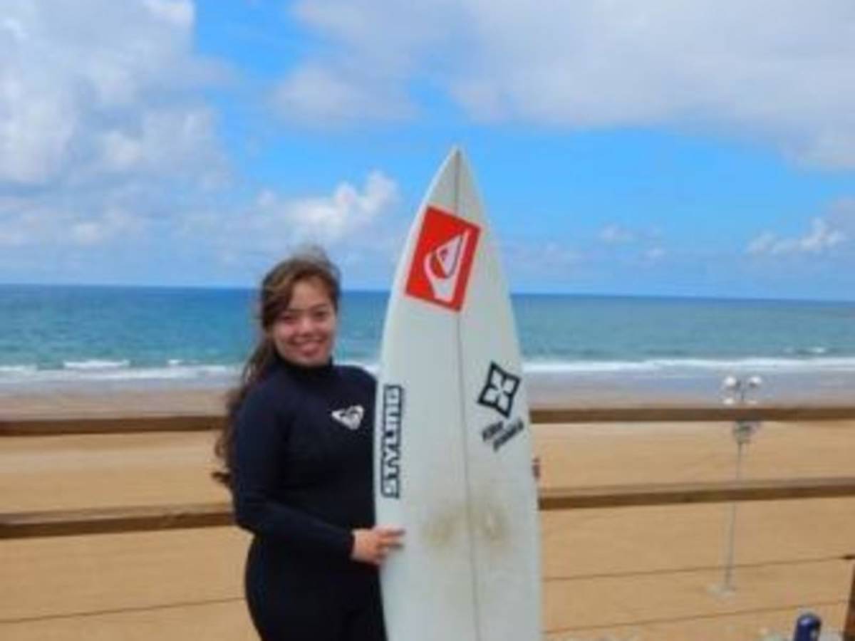 Person in a wetsuit holding a surfboard on a beach, with the ocean and a cloudy sky in the background.