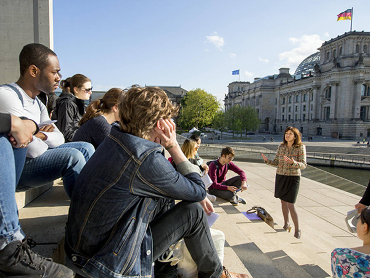 Seated students gathered on steps listen to an instructor. The class is outside across the river from the Reichstag.