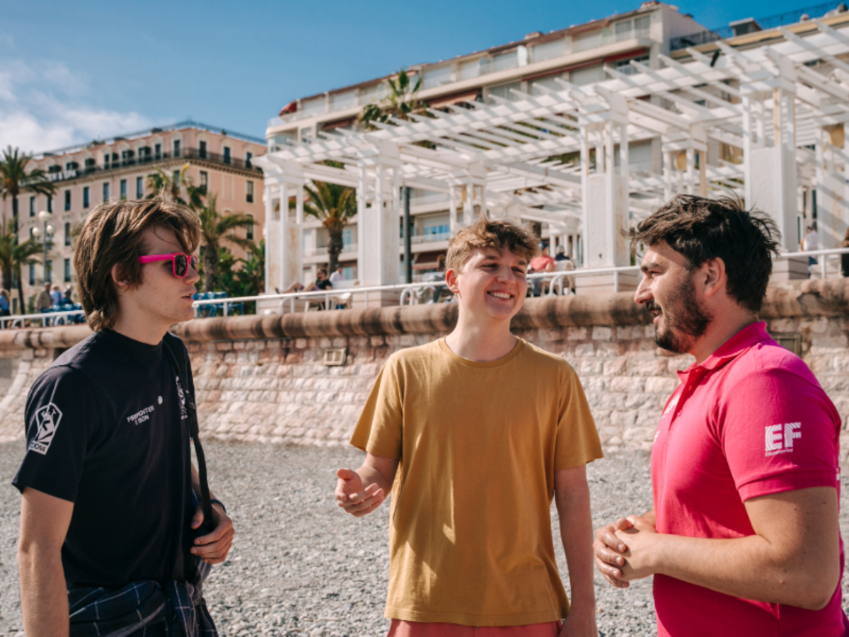 Students chatting with an EF staff member on the pebble beach in Nice, with palm trees and seaside architecture in the background.