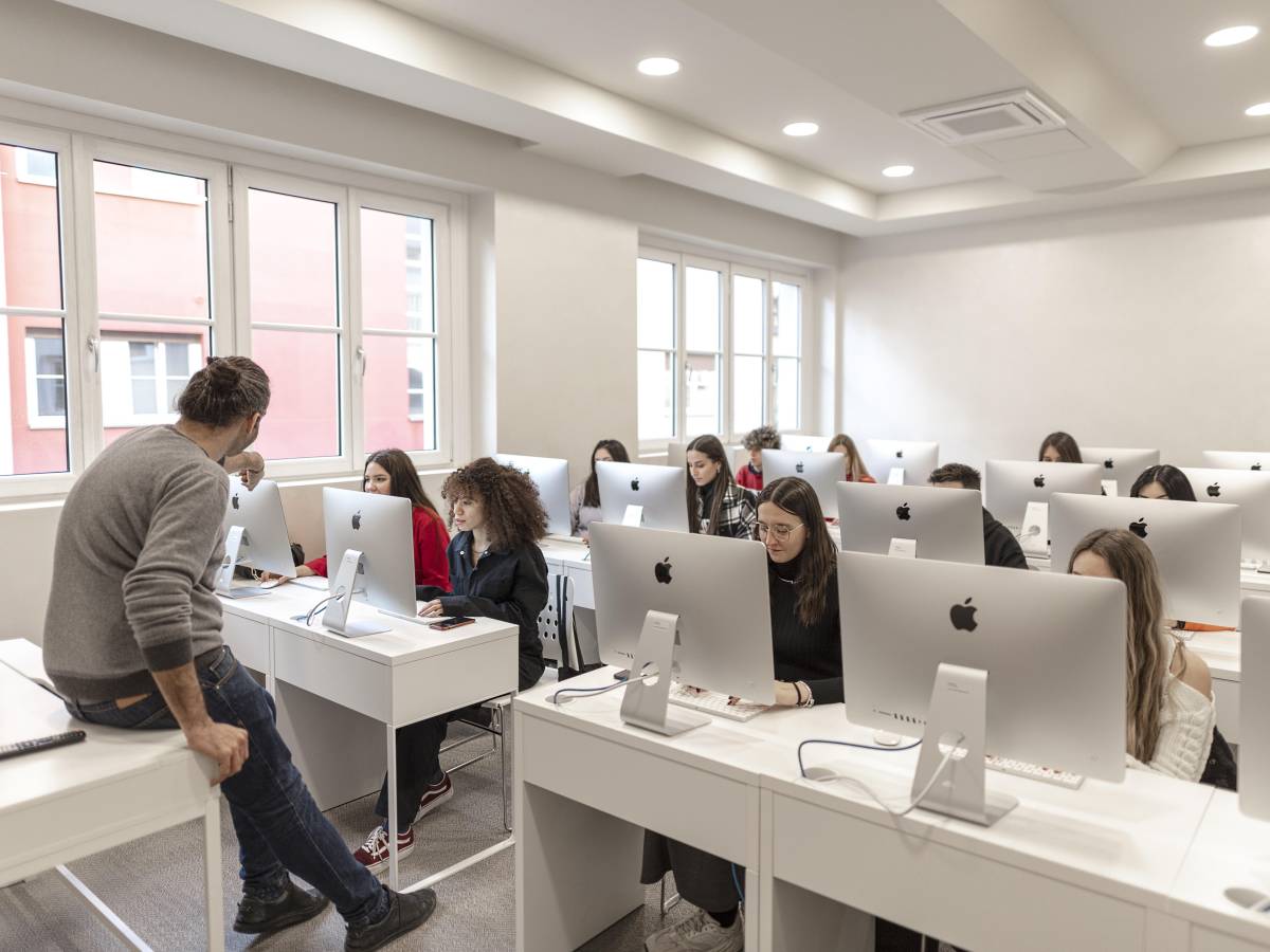 Students sat behind apple computers while a teacher sits on his desk in front of them