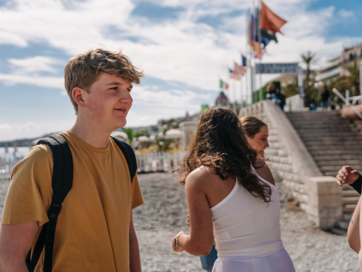 Students standing and chatting near the Promenade des Anglais in Nice, with flags and stone steps in the background.