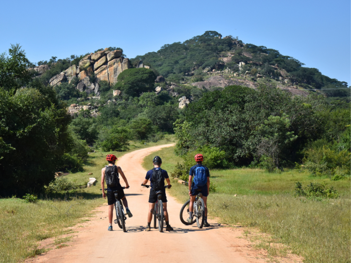 Three cyclists wearing helmets and backpacks stand on a dirt path, gazing at a rocky hill surrounded by lush greenery under a clear blue sky.