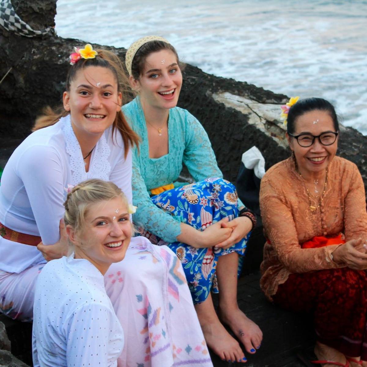 Three students and an Indonesian woman in traditional attire. They are sitting on a rock formation next to an ocean.