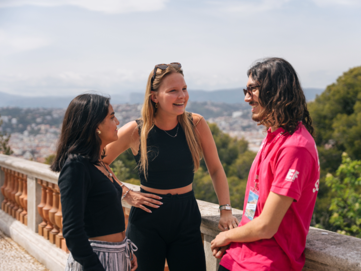 Three students talking and smiling on a scenic viewpoint in Nice, with the city and hills in the background.