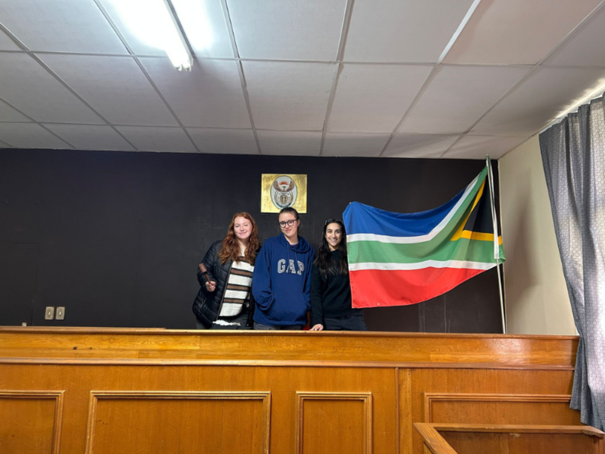 Three volunteers stand in a mock courtroom, next to the flag of South Africa, smiling into the camera. 