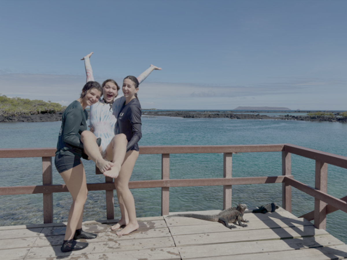 Three women pose playfully on a wooden pier near water.