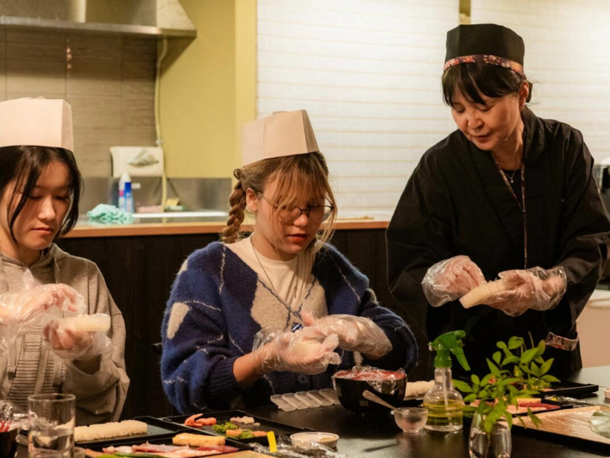 Two EF Gap Year students in Tokyo making sushi with guidance from a Japanese chef, wearing gloves and hats while participating in a cultural cooking class.