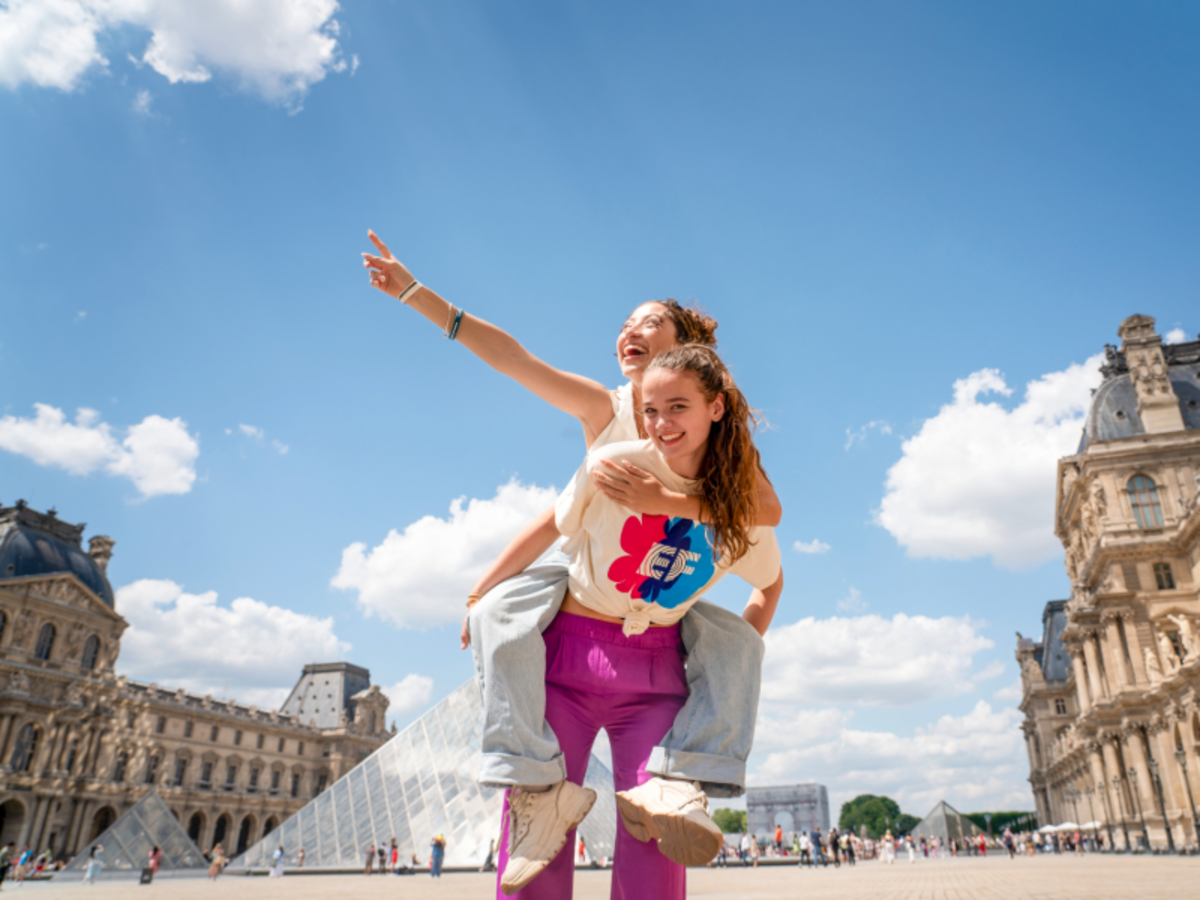 Two EF students enjoying a playful piggyback ride in front of the Louvre Pyramid in Paris on a sunny day.