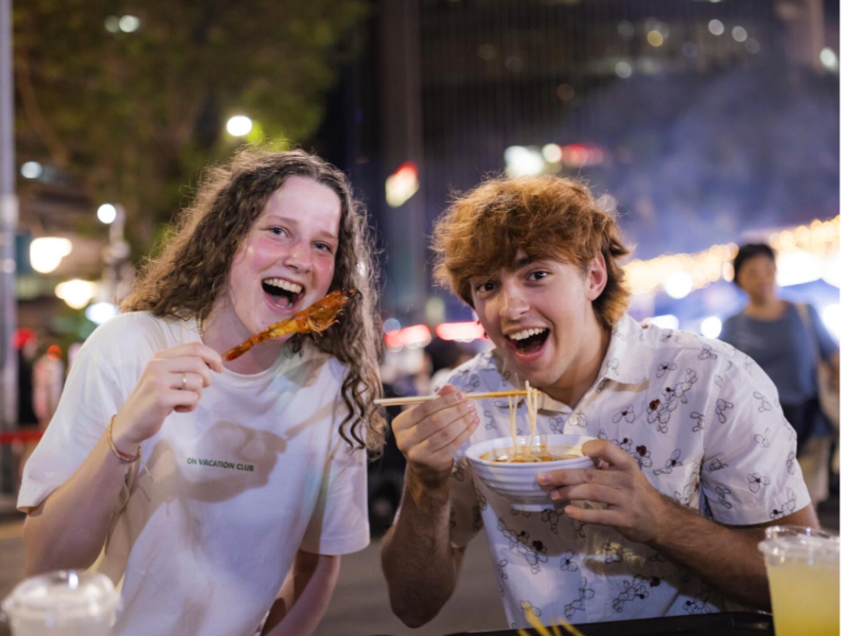 Two EF students enjoying local street food at night, smiling while trying skewers and noodles.