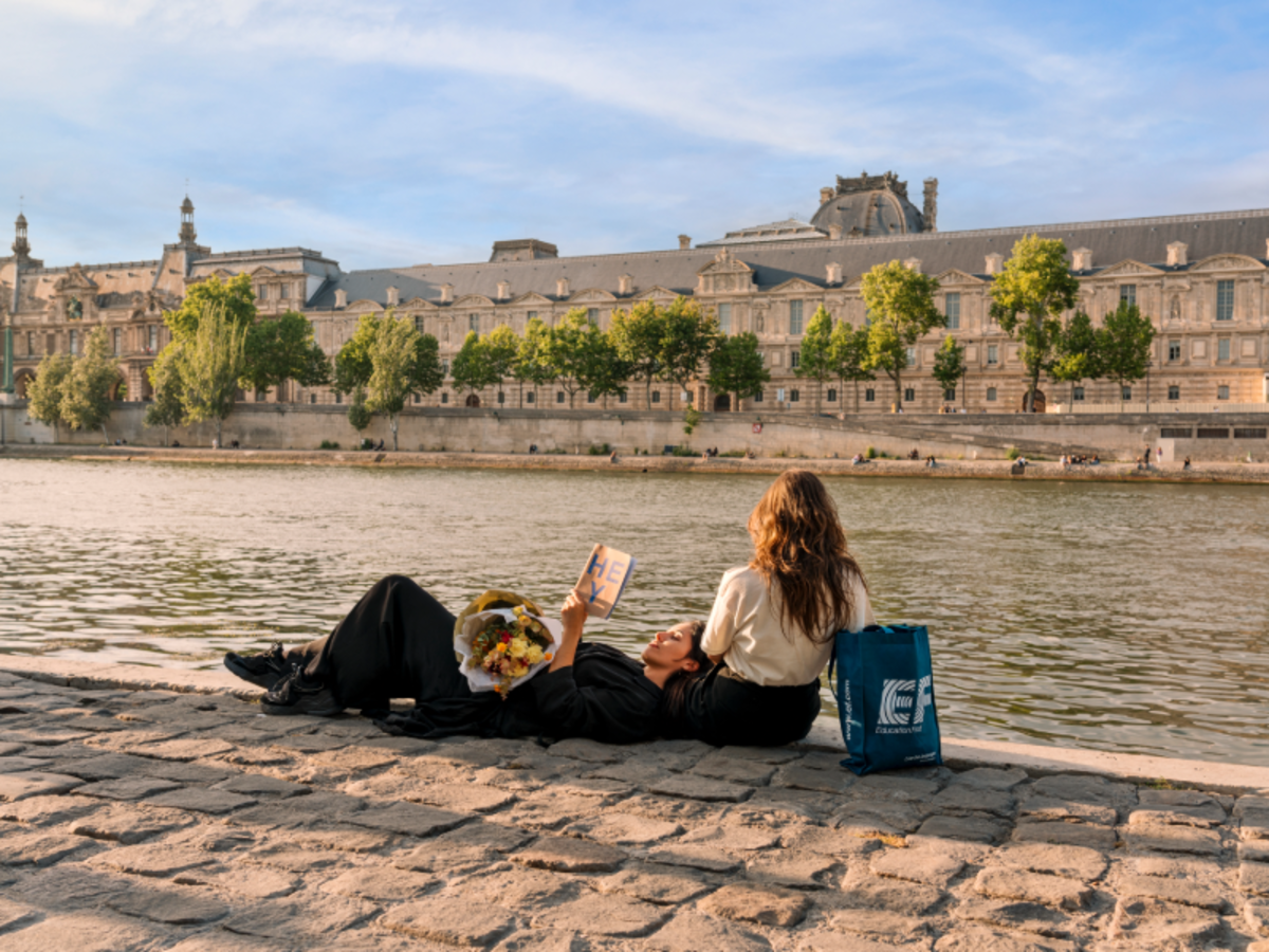 Two EF students relaxing by the Seine River in Paris, one holding flowers and a book, with the Louvre visible in the background.