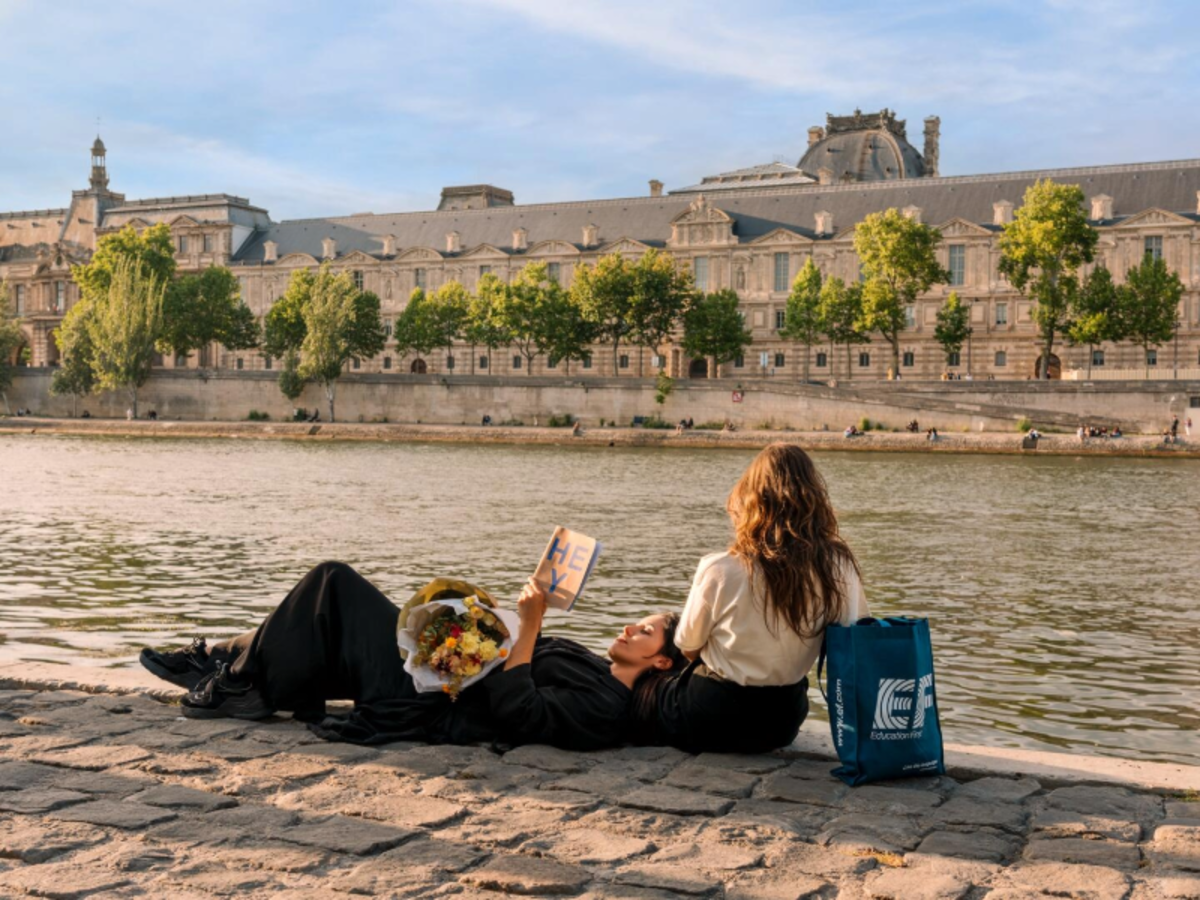 Two EF students relaxing by the Seine River in Paris, with flowers, a book, and an EF tote bag beside them.