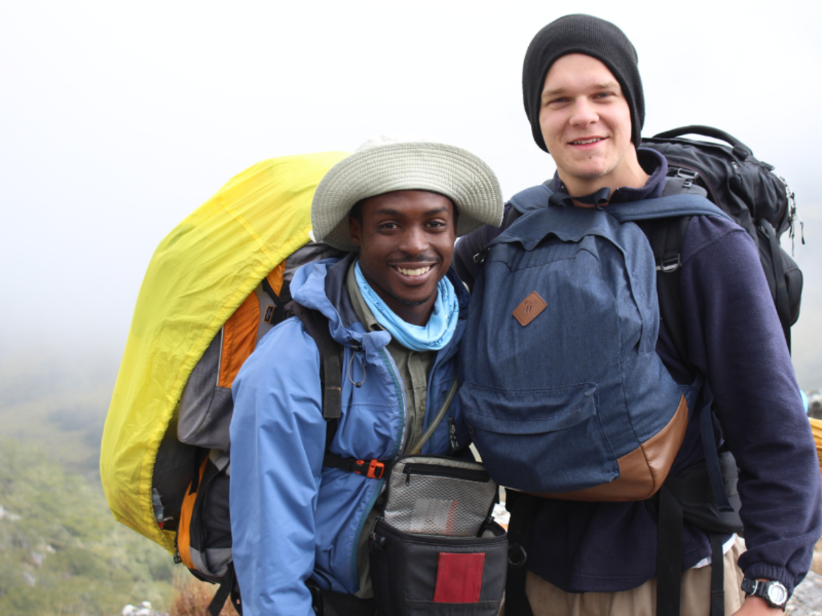 Two hikers with large backpacks, smiling in a misty, mountainous landscape. One wears a sun hat and blue jacket, the other a beanie and navy jacket.