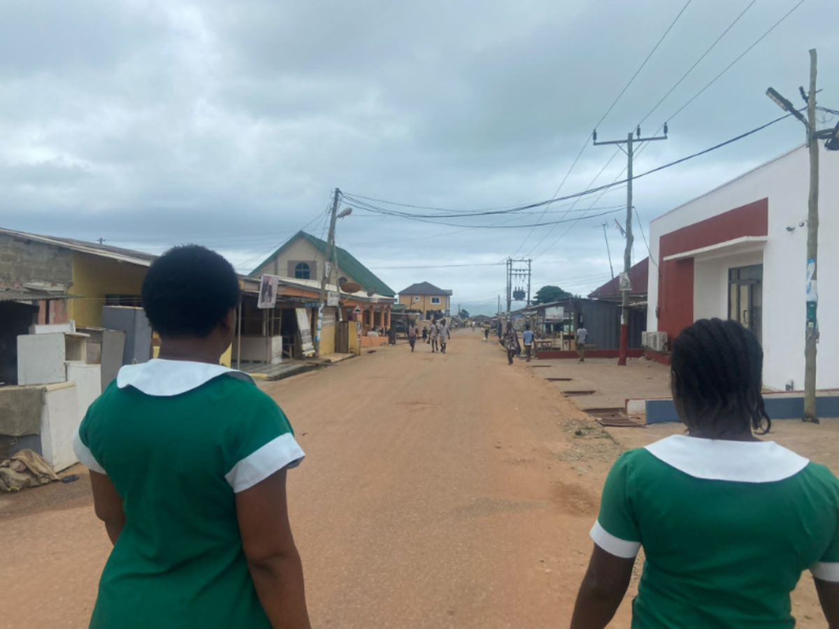 Two nurses in green and white shirts walk away from the camera down an orange dust road with small buildings either side.