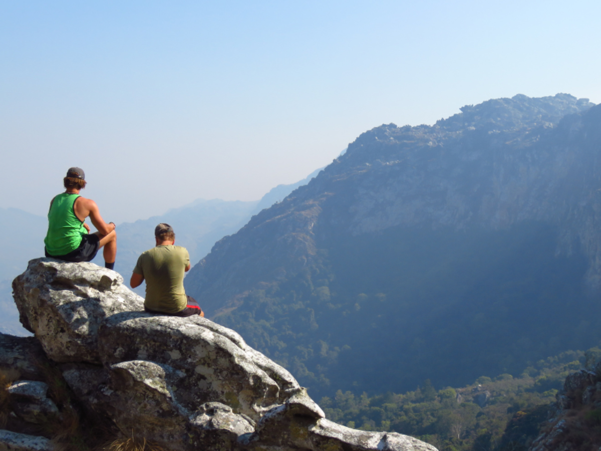 Two people sit on rocky ledge, overlooking vast mountain landscape under clear blue sky. The scene conveys tranquility and awe.