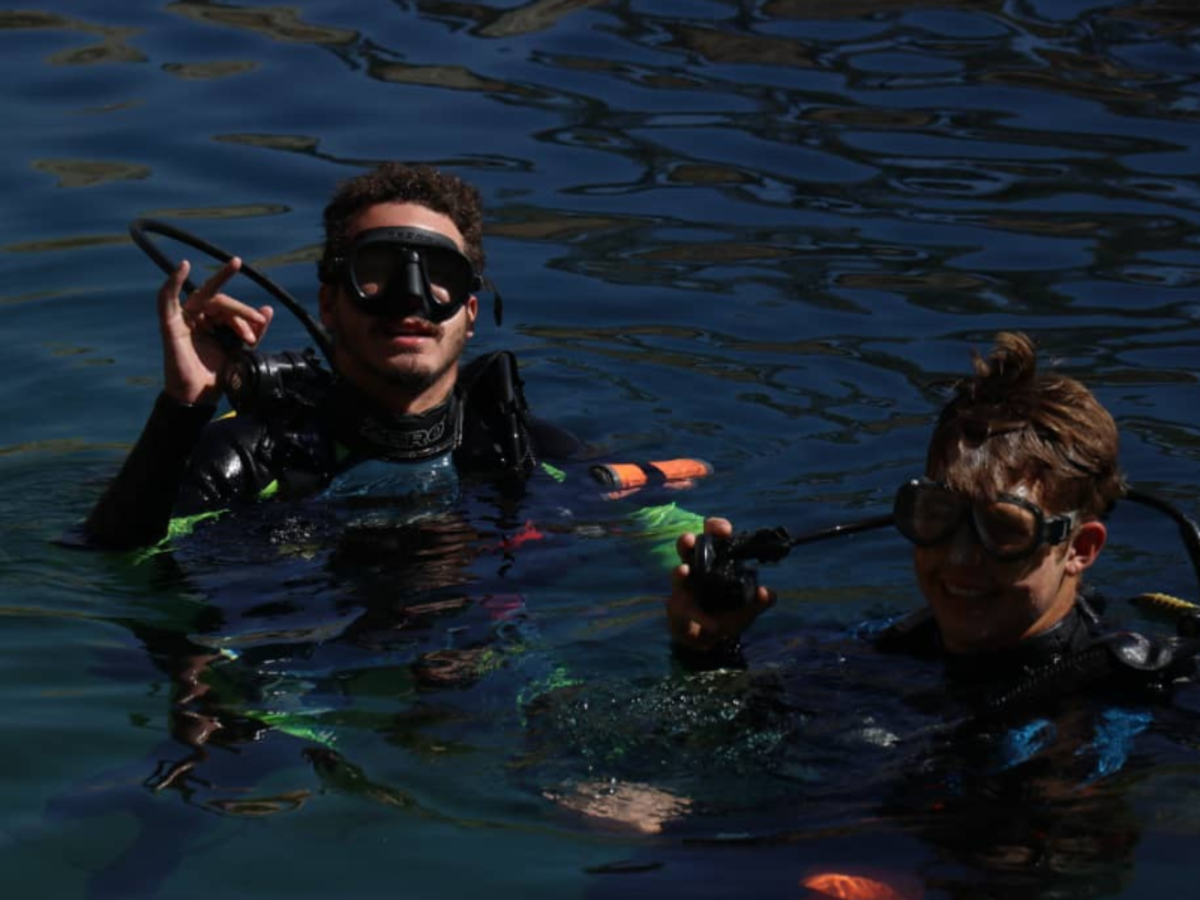 Two scuba divers are in dark blue water, wearing black masks and wetsuits. One gives an OK sign, both smiling, conveying a sense of excitement and adventure.