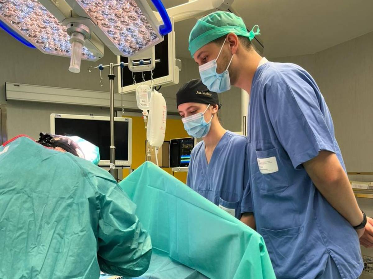 Two students dressed in scrubs are peering over an operating table, witnessing a surgery.