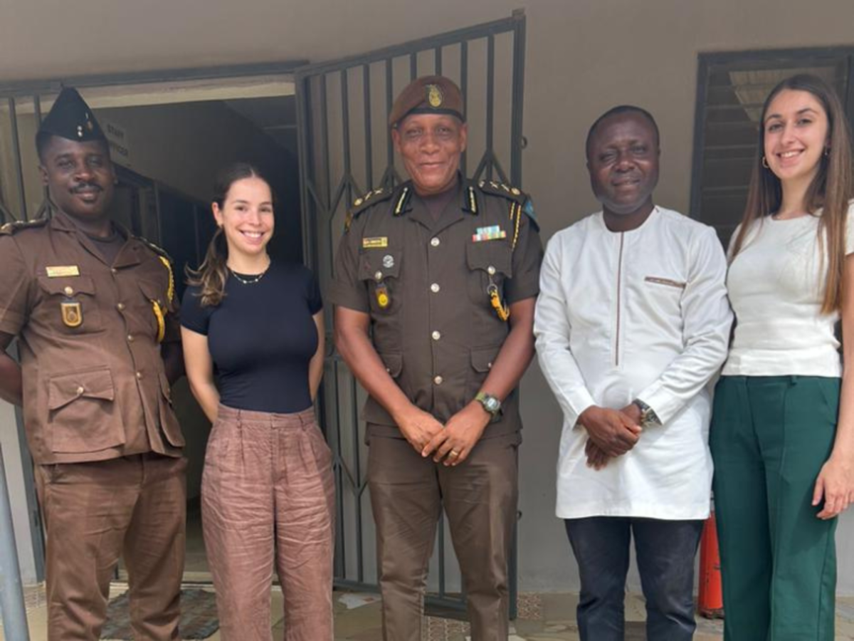 Two volunteers stand smiling with their programme coordinator, as well as two prison officials who don brown berets and brown uniforms. 