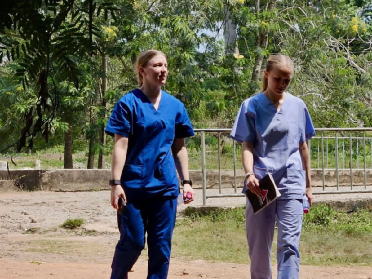 Two volunteers wearing blue scrubs walk through the trees to their placement.