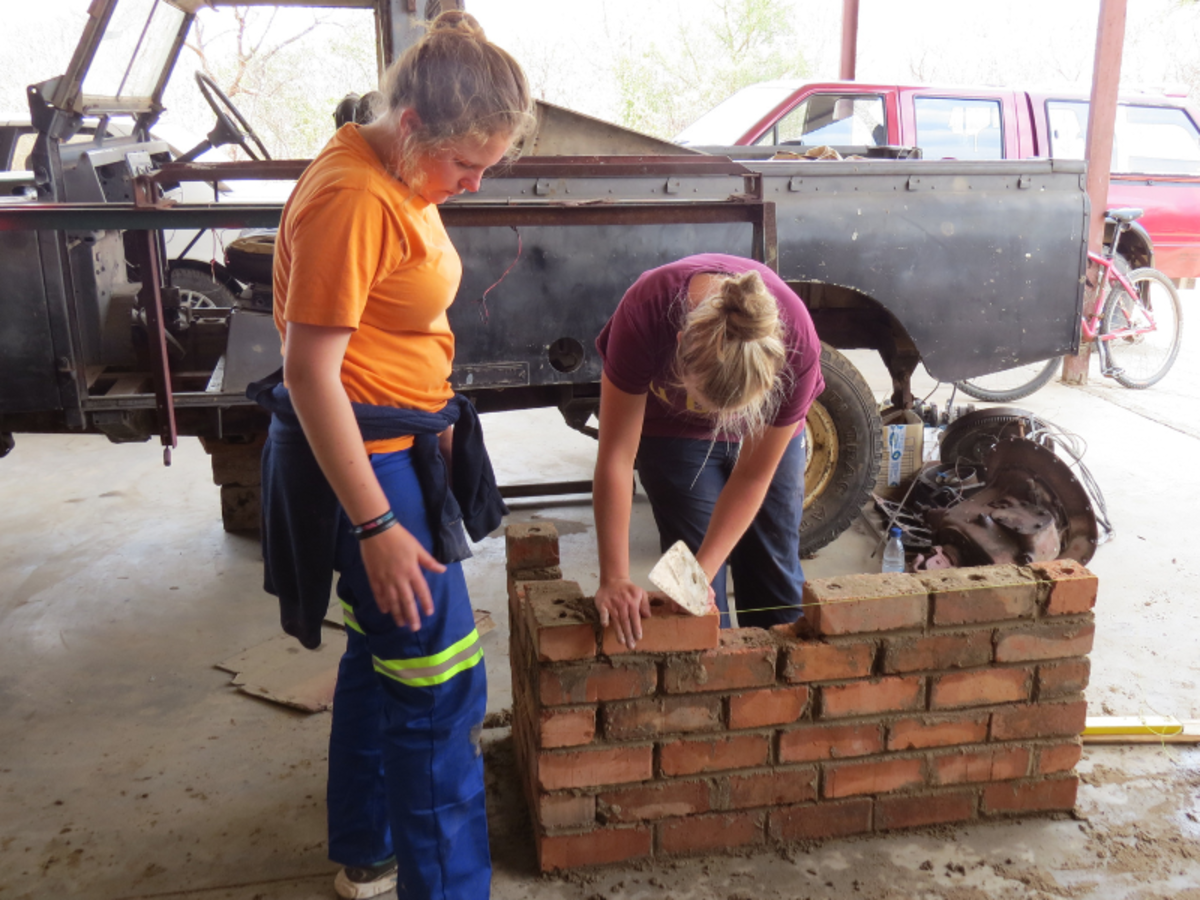Two women build a brick wall in a workshop, surrounded by car parts. One lays a brick while the other observes, conveying focus and teamwork.