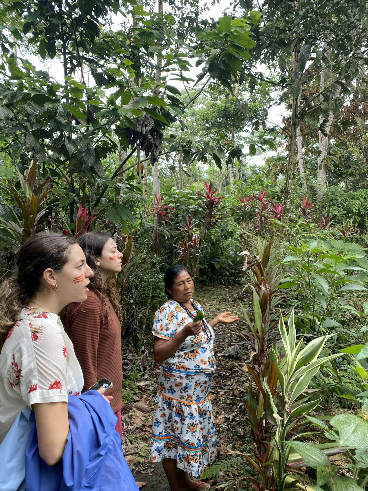 Two women listen to a Kichwa woman speak in a garden setting. 