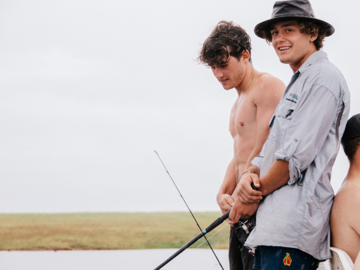 Two young men fishing by a lake. One is shirtless holding a rod, the other in a hat and casual shirt smiles. The scene is relaxed under an overcast sky.