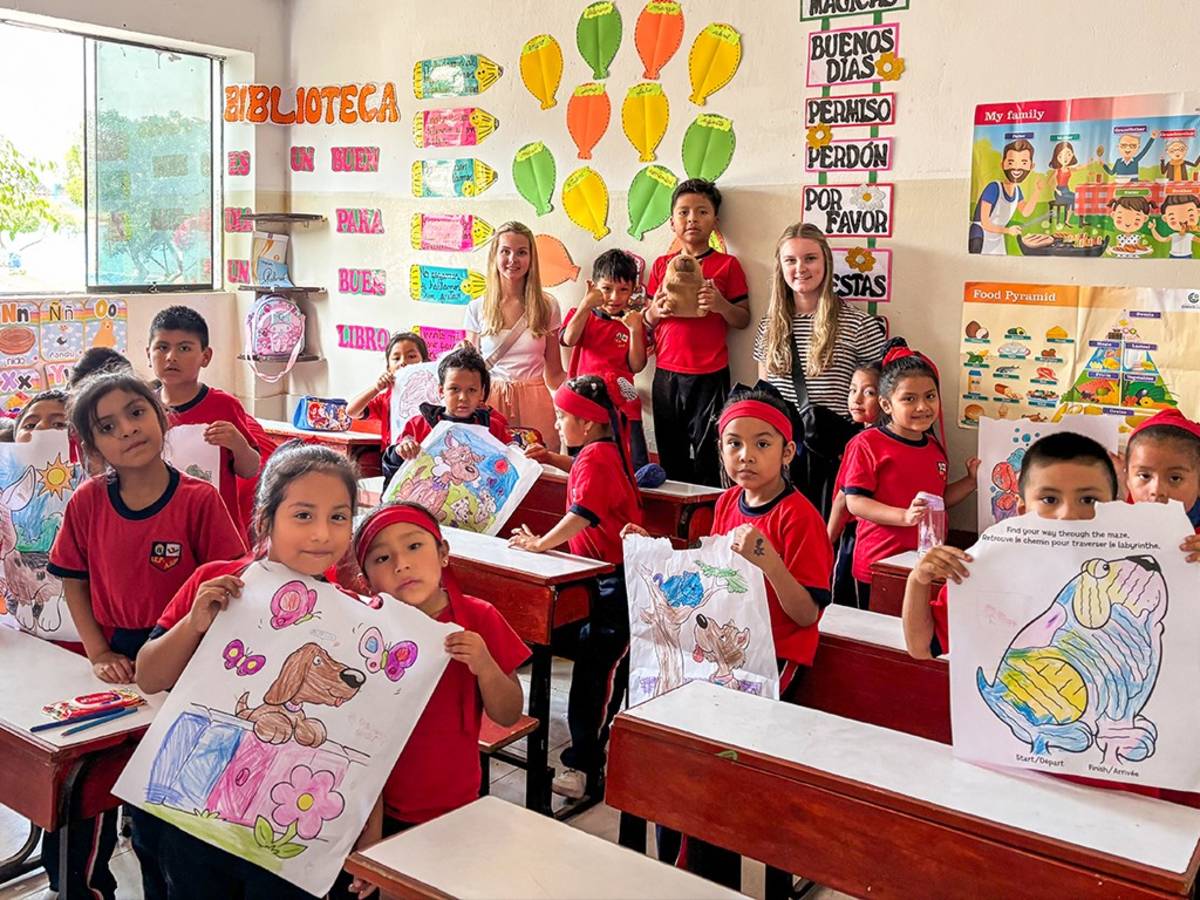Two young volunteers stand with a classroom of Peruvian students in red uniforms proudly displaying their colorful drawings.