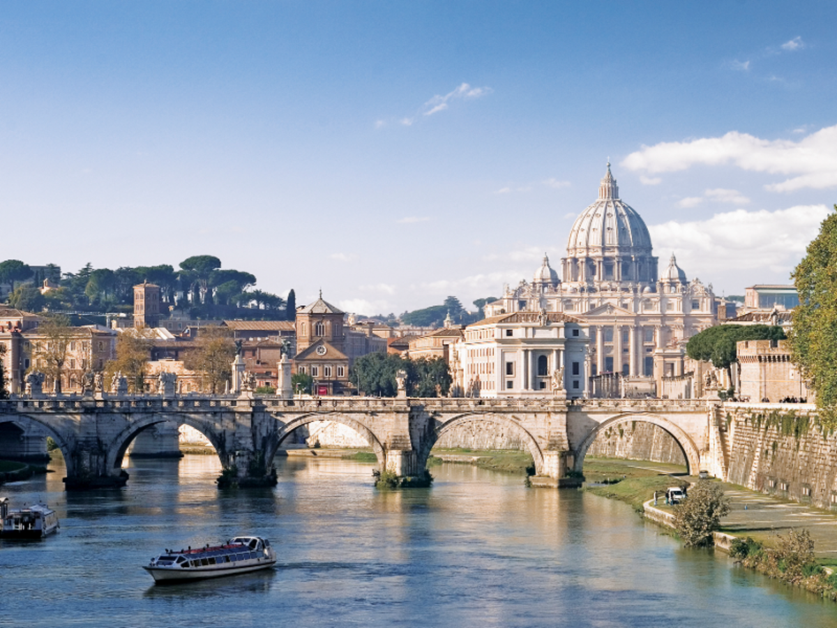 View of St. Peter’s Basilica and the Vatican across the Tiber River in Rome, with boats passing under the historic stone bridge.