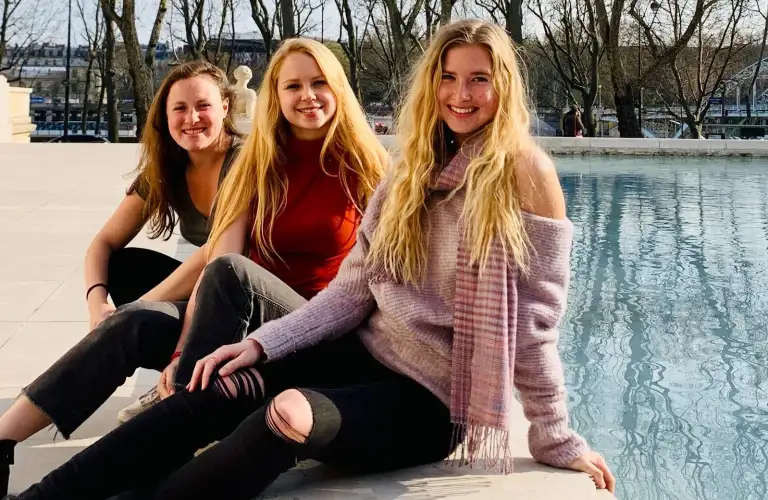 Three women smiling by a pool with Eiffel Tower in the background.