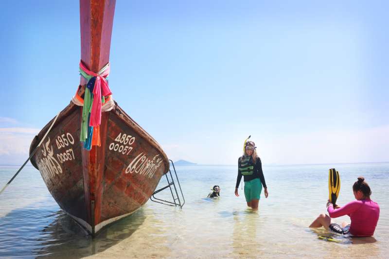 A decorative longtail boat with Thai script is resting on the shore. 3 students are putting on snorkelling gear. The water is crystal clear and the sand is white, with blue sky.