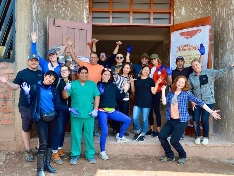 A diverse group of cheerful volunteers and staff pose together outside a community building in Peru, many wearing blue gloves and waving.