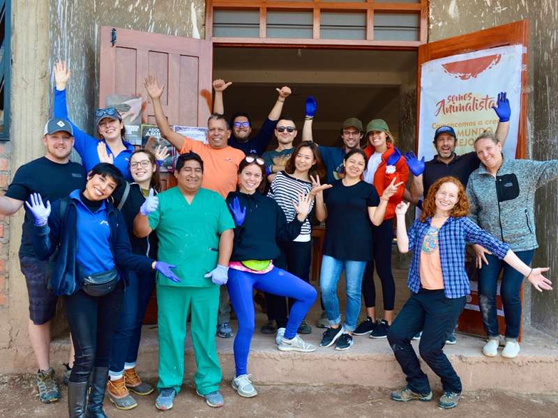 A diverse group of cheerful volunteers and staff pose together outside a community building in Peru, many wearing blue gloves and waving.