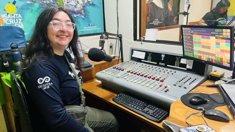 A girl poses in front of a microphone and computer setup in an office setting.
