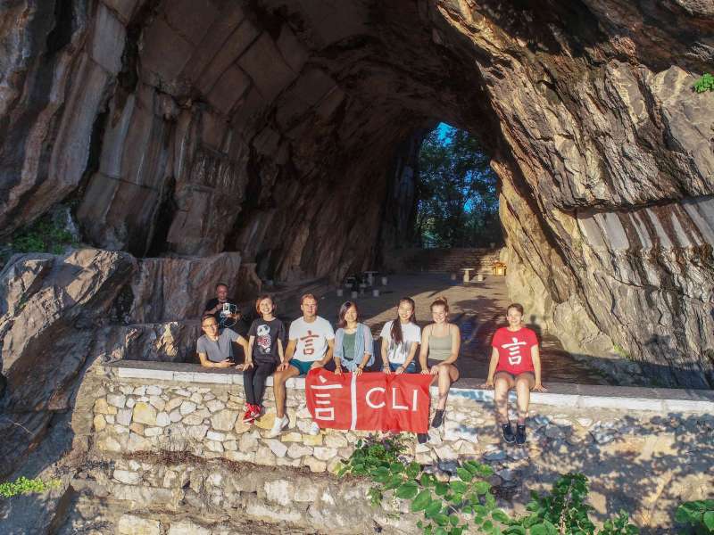A group of CLI students and teachers sit on a stone wall holding a red CLI flag at the entrance of a large karst cave in Guilin.
