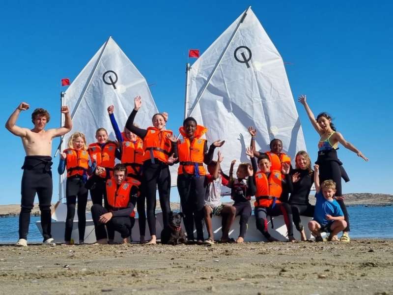 A group of interns and students in wetsuits and orange life jackets pose cheerfully on the beach in front of two white sailing dinghies.