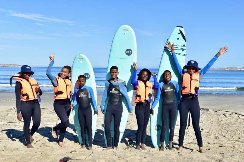 A group of Lüderitz Blue School boarding students in wetsuits and life jackets posing with surfboards on the beach at the South Atlantic Ocean.