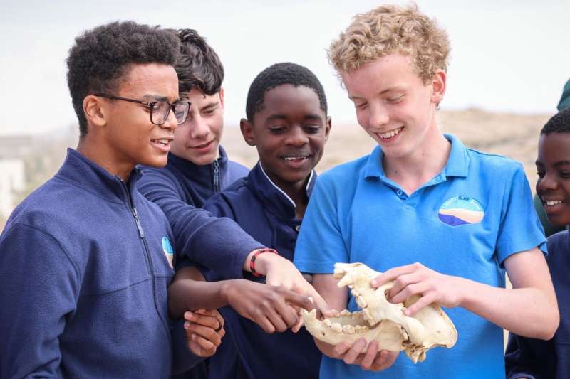 A group of Lüderitz Blue School students in uniform examining a specimen together outdoors during an Ocean Day visit from the Brown Hyena Research Project.