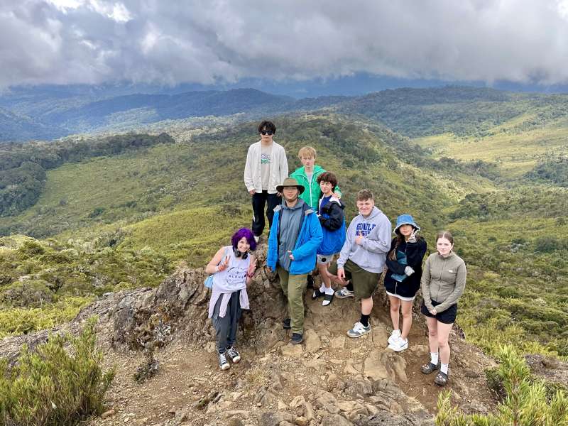 A group picture posed on top of a mountain, with green hills in background.