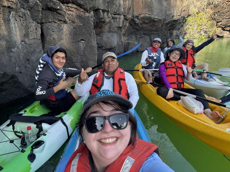 A group poses for a selfie, all in kayaks in the water with a rock wall behind them.
