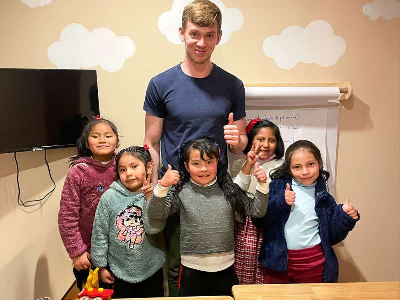 A male volunteer and five young Peruvian girls smile and give thumbs-up in a classroom with cloud wall decals in Cusco.